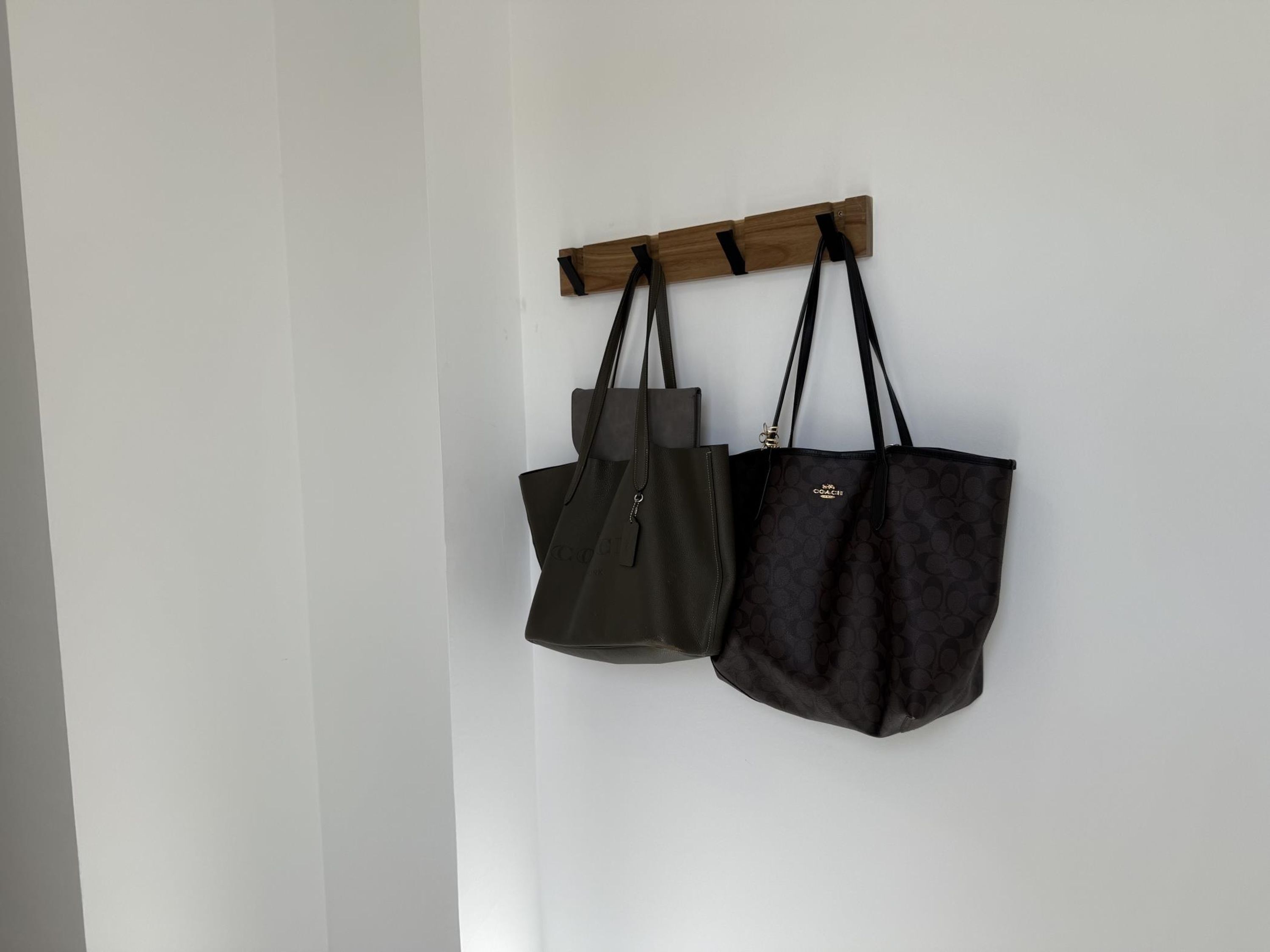 Deux sacs suspendus à un porte-manteau en bois, le tout adossé à un mur blanc.