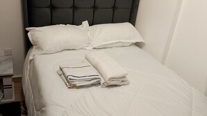 Un lit bien fait, avec du linge de lit blanc, une serviette pliée et une petite table de chevet surmontée d’une lampe.