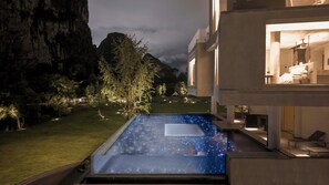 Un edificio moderno con una piscina infinita de vidrio por la noche, rodeado de un jardín bien iluminado y montañas al fondo.