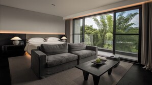 A modern hotel room with a large bed, a grey sofa, a small table with a fruit bowl, and a large window offering a view of trees. - YINGMAN HOTEL (Guilin)