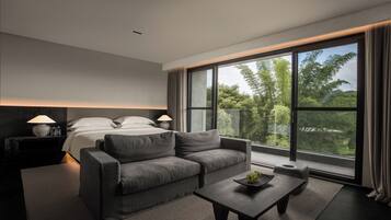 Une chambre d’hôtel moderne avec un grand lit, un canapé gris, une petite table sur laquelle se trouve un panier de fruits, et une grande fenêtre donnant sur des arbres.