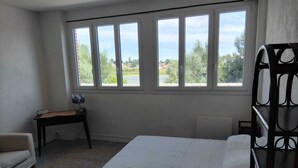 Un dormitorio con cama, escritorio y silla. Amplias ventanas ofrecen vistas al exterior.