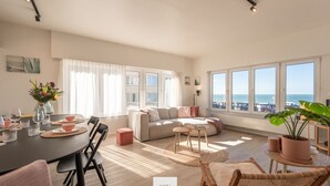 A spacious living area with a large window overlooking the sea, a dining table set with glasses and a vase of flowers, and a cozy seating area with a sofa and chairs. - The Corner Floor, front row sea view and parking (Oostende)