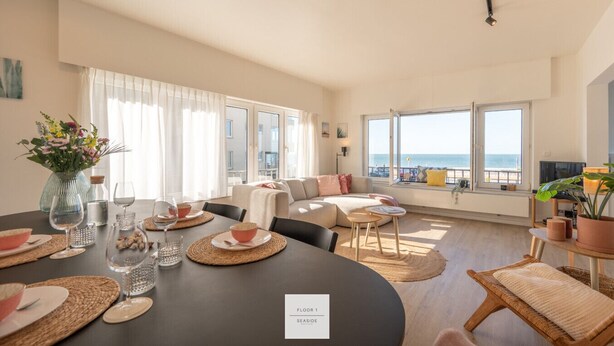 A spacious living room with a large window overlooking the sea, a dining area with a table set for two, and a cozy seating area with a sofa and cushions. - The Corner Floor, front row sea view and parking (Oostende)