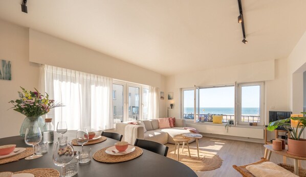 A spacious living room with a large window overlooking the sea, a dining area with a table set for two, and a cozy seating area with a sofa and cushions. - The Corner Floor, front row sea view and parking (Oostende)