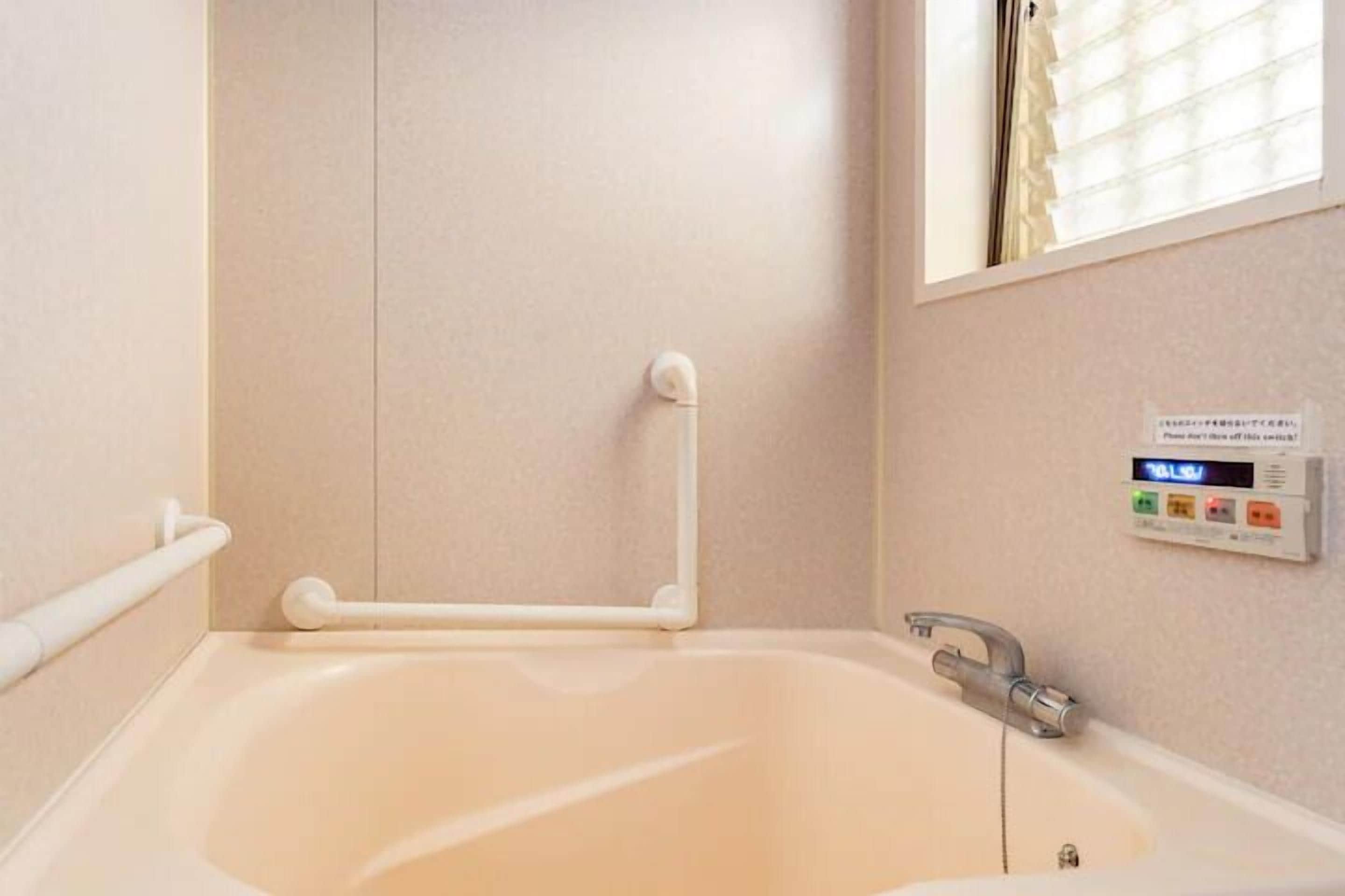 A bathroom with a bathtub, grab bars, a digital thermostat, and a window with blinds.