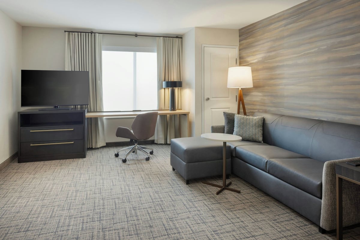 A hotel room with a flat-screen TV, a desk with a lamp, a grey sofa, and a small round table.