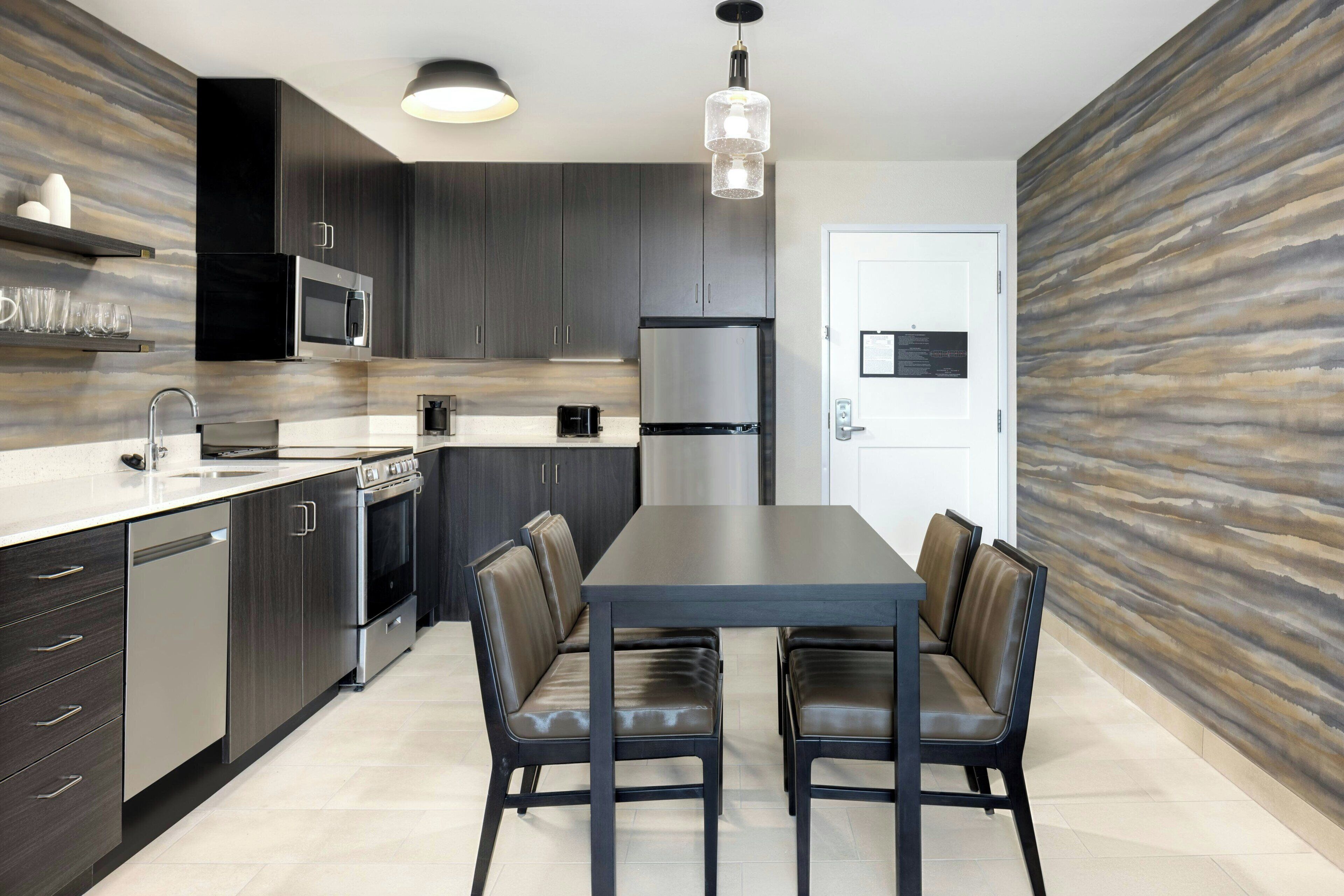 A modern kitchen with dark cabinetry, stainless steel appliances, a dining table with leather chairs, and a textured wall.