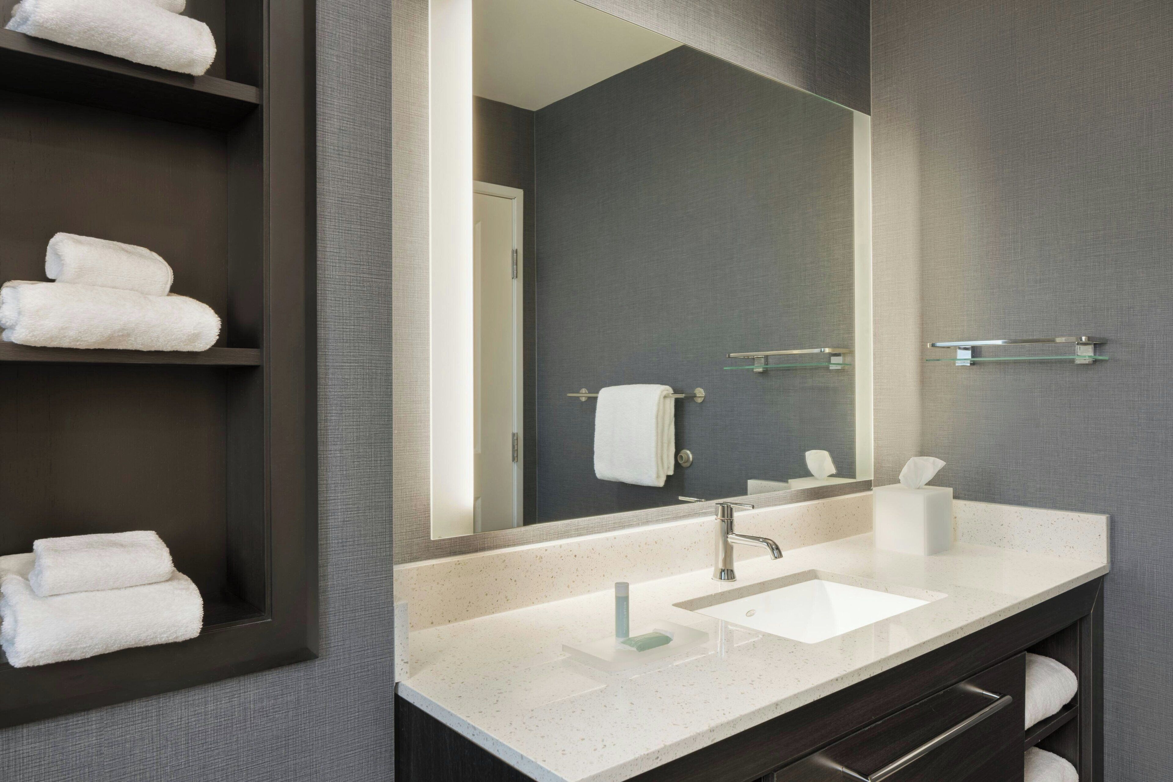 Un baño moderno con un espejo grande, tocador de madera oscura, lavamanos blanco y varias toallas en estantes.