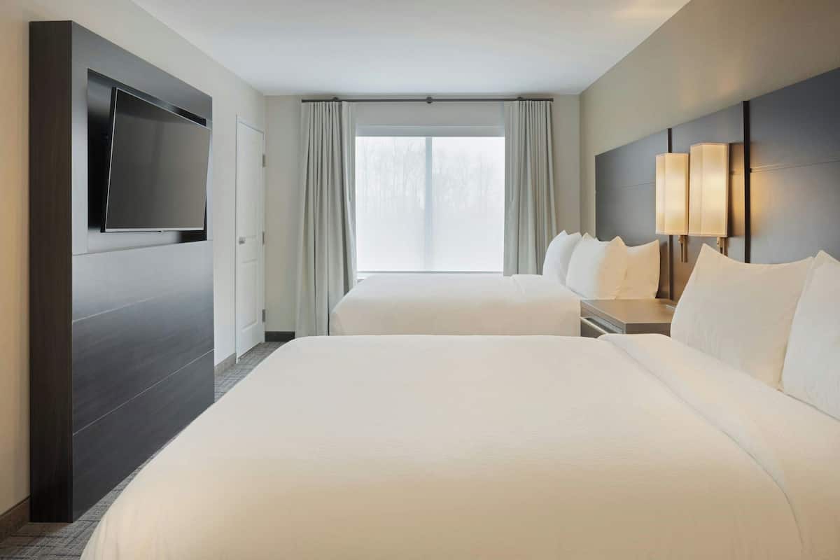 A hotel room with a large bed, a TV mounted on the wall, a bedside table with lamps, and a window with curtains.
