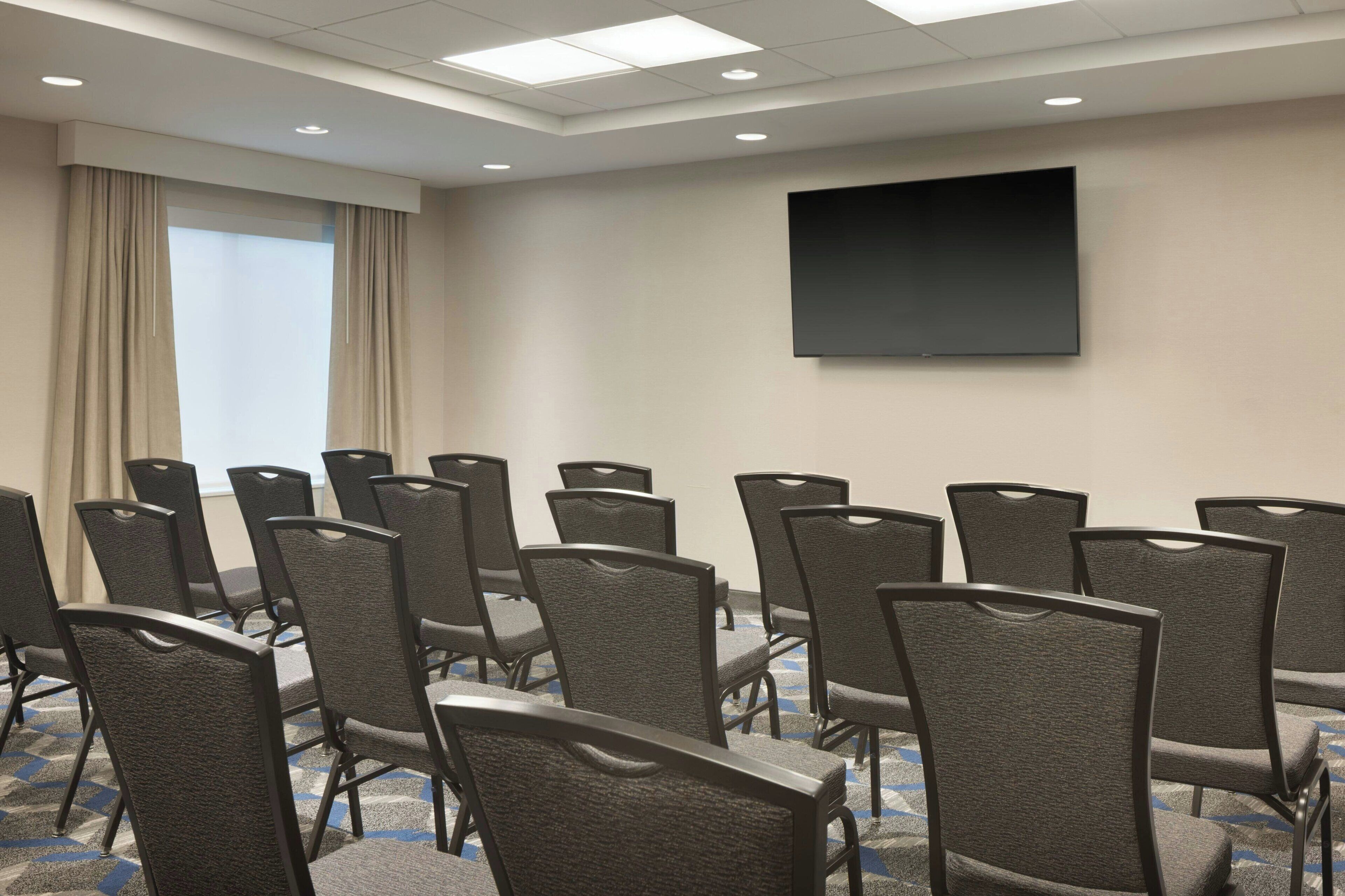 Un salón de conferencias con filas de sillas, una pantalla grande y un ventanal con cortinas.