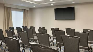 Un salón de conferencias con filas de sillas, una pantalla grande y un ventanal con cortinas.