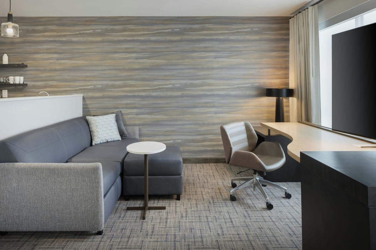 A modern hotel room with a gray sofa, a small round table, a desk with a chair, and a large window with curtains.