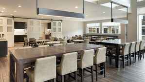 A spacious dining area with a long wooden table and multiple chairs, a kitchen in the background, and large windows providing natural light. - Residence Inn by Marriott Battle Creek (Battle Creek)
