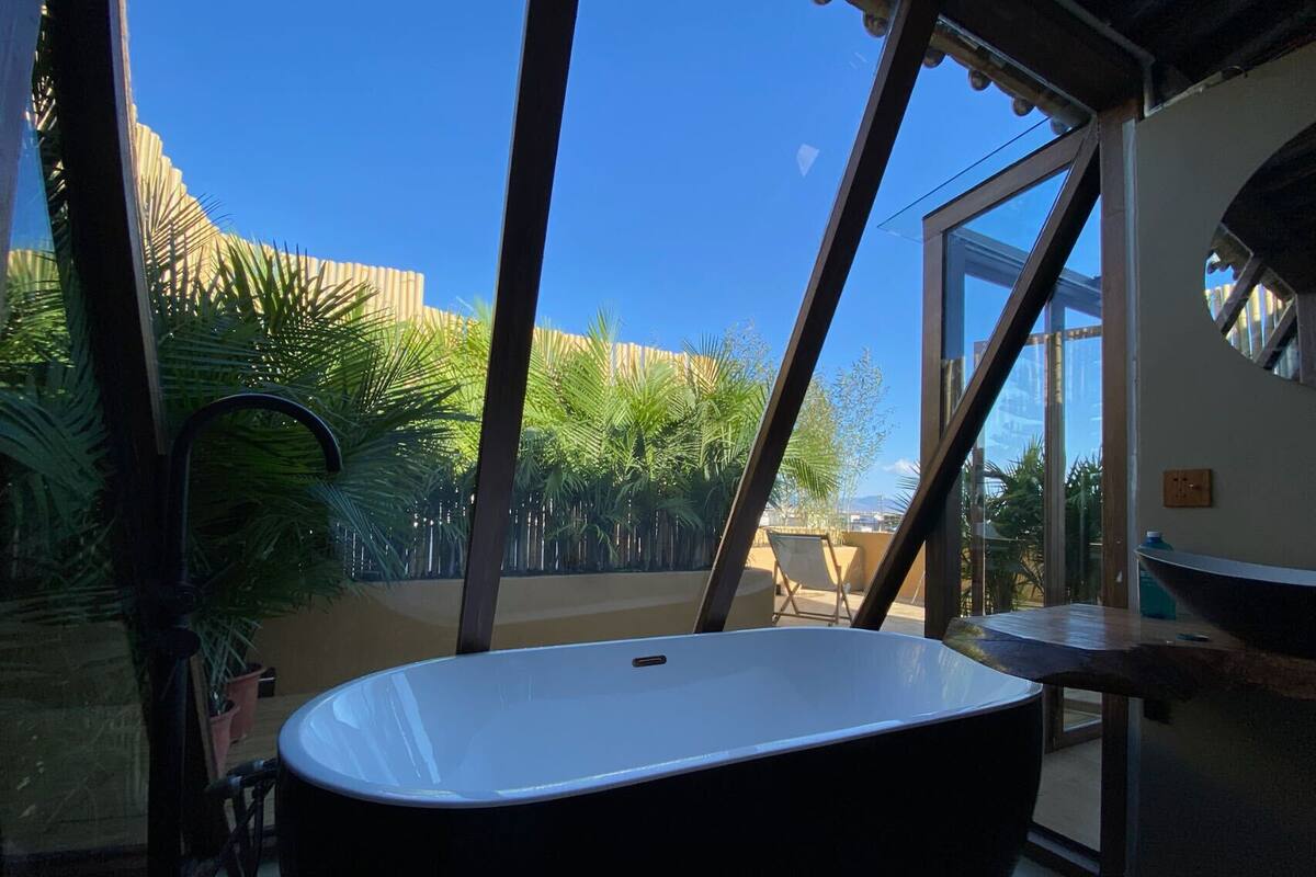 A modern bathroom with a freestanding bathtub, a large window, and a view of a palm tree garden.
