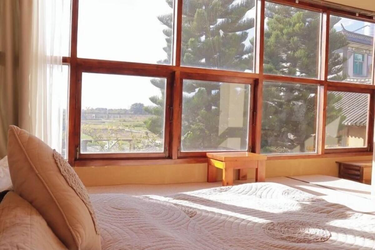 A room with a large window, a bed, a wooden bench, and a small table.