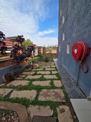A rooftop garden with a stone pathway, a red fire hose, and a blue building in the background. - Golden Flame (Kigali)