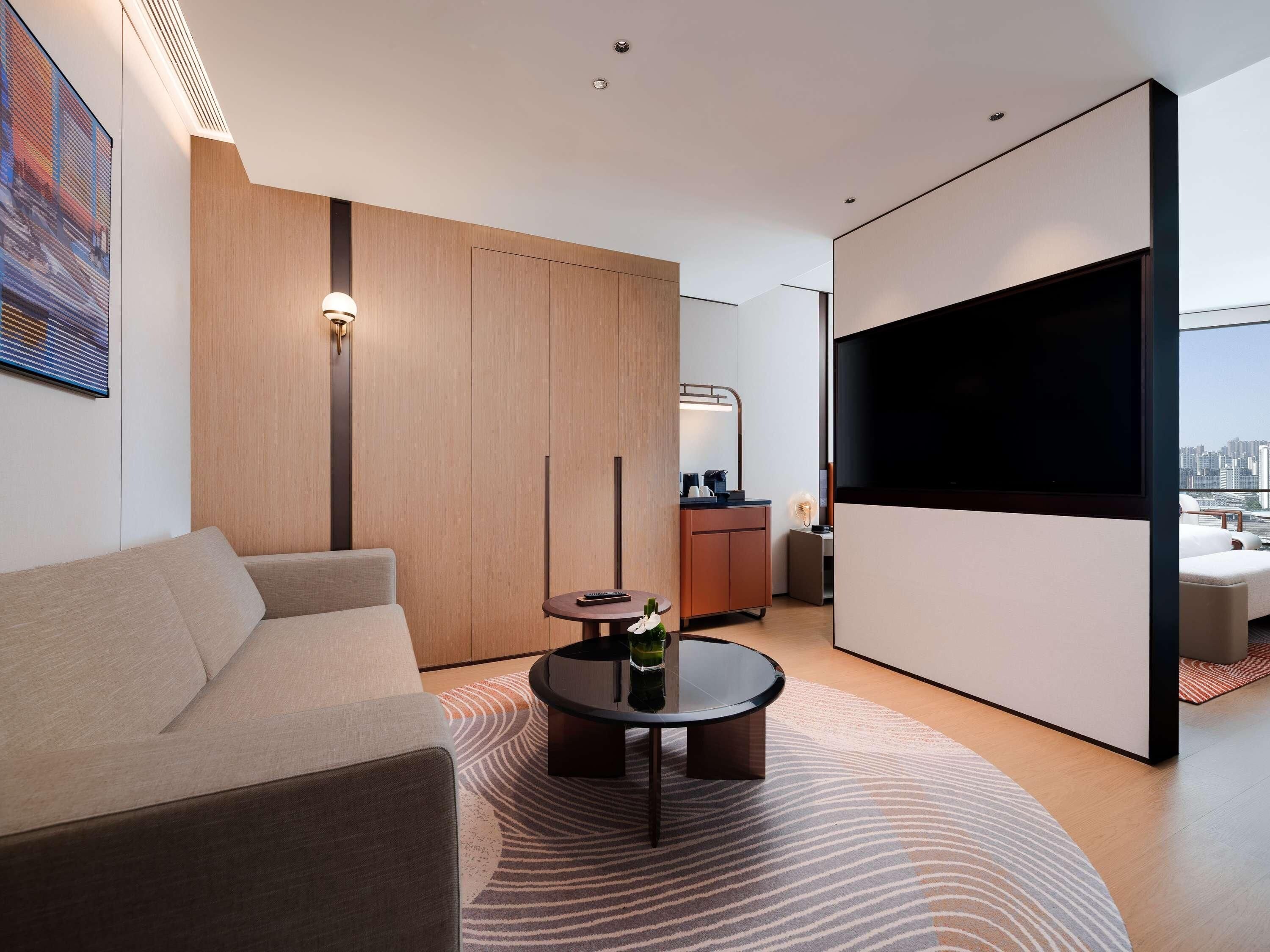 A modern hotel room with a sofa, a round coffee table, a TV, and a city view.
