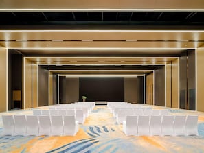 Un salón de conferencias con filas de sillas blancas, piso alfombrado y una pantalla grande al frente.