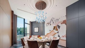 Een moderne eetkamer met een ronde tafel gedekt voor zes personen, omringd door donkerbruine gestoffeerde stoelen. Een groot abstract kunstwerk aan de muur en een kroonluchter met blauwe glaselementen zijn zichtbaar.
