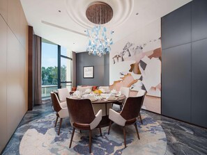 Een moderne eetkamer met een ronde tafel gedekt voor zes personen, omringd door donkerbruine gestoffeerde stoelen. Een groot abstract kunstwerk aan de muur en een kroonluchter met blauwe glaselementen zijn zichtbaar.