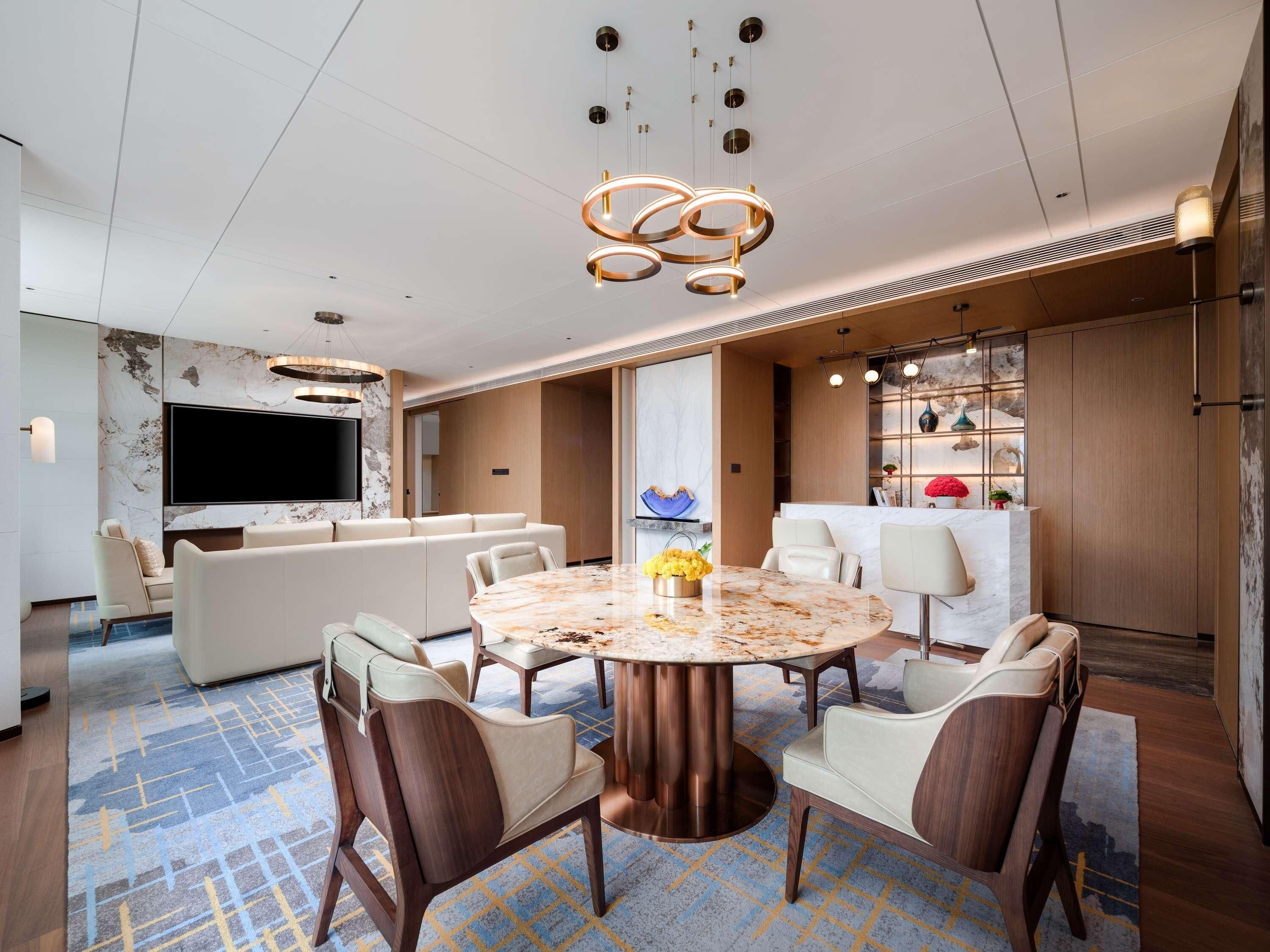Quarto de hotel moderno com uma mesa de jantar redonda de mármore, assentos confortáveis, uma TV de tela plana e uma parede decorativa com prateleiras.