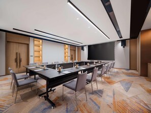 Een vergaderzaal met een lange tafel, stoelen, een scherm voor een beamer en een vloerbedekking.