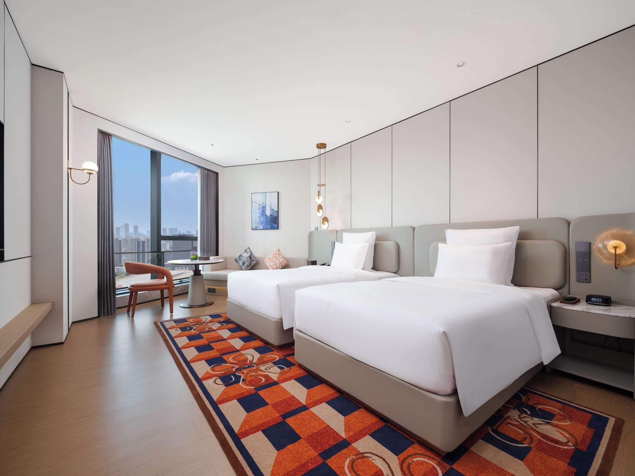 A modern hotel room with two beds, a desk, a chair, and a large window offering a city view.