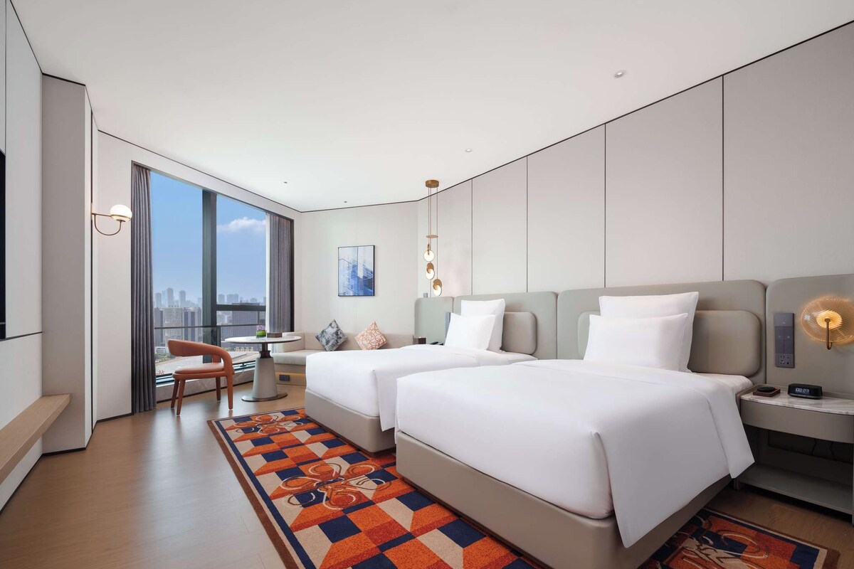 Een moderne hotelkamer met twee bedden, een bureau, een stoel en een groot raam met uitzicht op de stad.