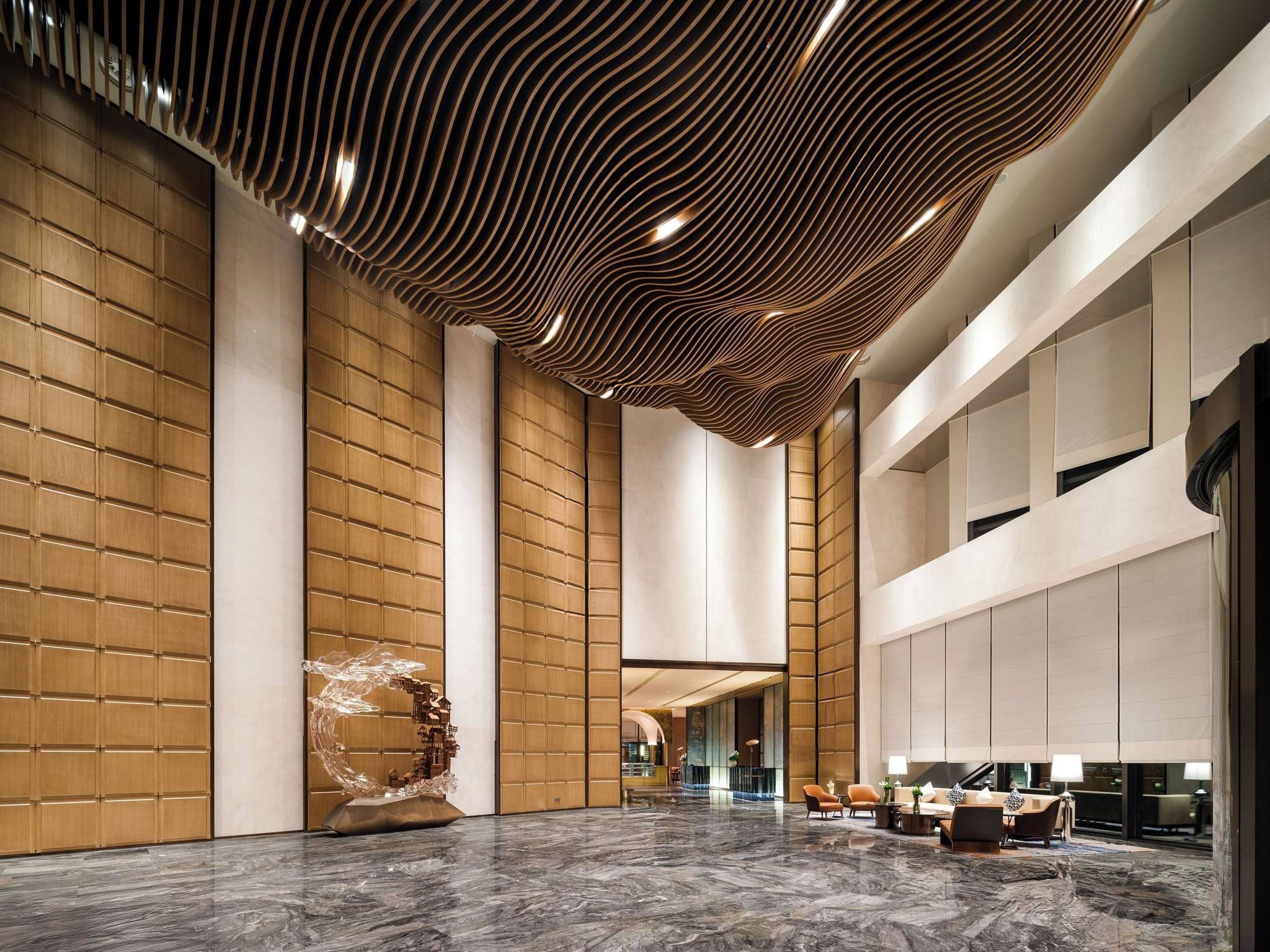 A spacious lobby with a high ceiling, wooden panel walls, a marble floor, and a seating area with sofas and chairs.