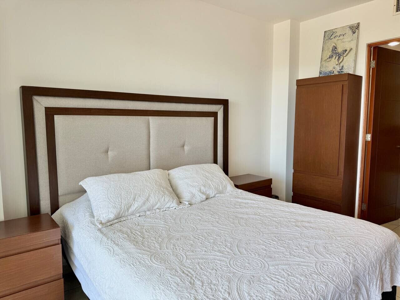 Una cama bien hecha con una colcha blanca y dos almohadas, una mesita de noche de madera con cajón y un armario de madera frente a una pared lisa.