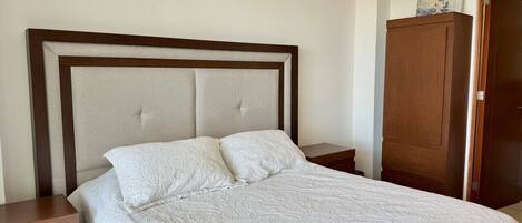Una cama bien hecha con una colcha blanca y dos almohadas, una mesita de noche de madera con cajón y un armario de madera frente a una pared lisa.