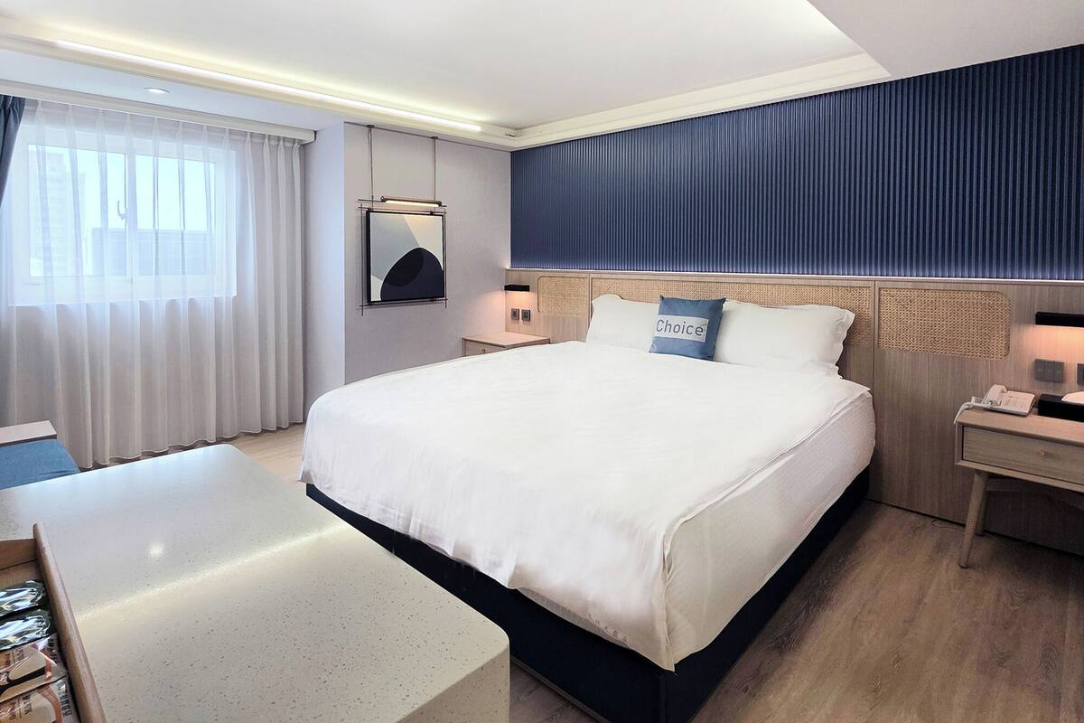 Habitación de hotel con una cama grande, un escritorio, una silla, una mesita de noche y una ventana con cortinas.