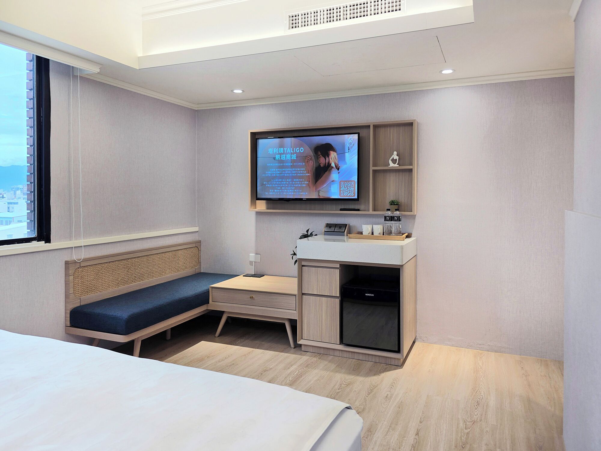 A modern hotel room with a bed, a seating area, a TV, a small desk, and a window.
