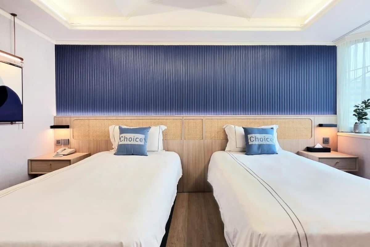 Una habitación de hotel con dos camas, un ventanal grande, un panel de pared azul y una mesita de noche con lámpara.