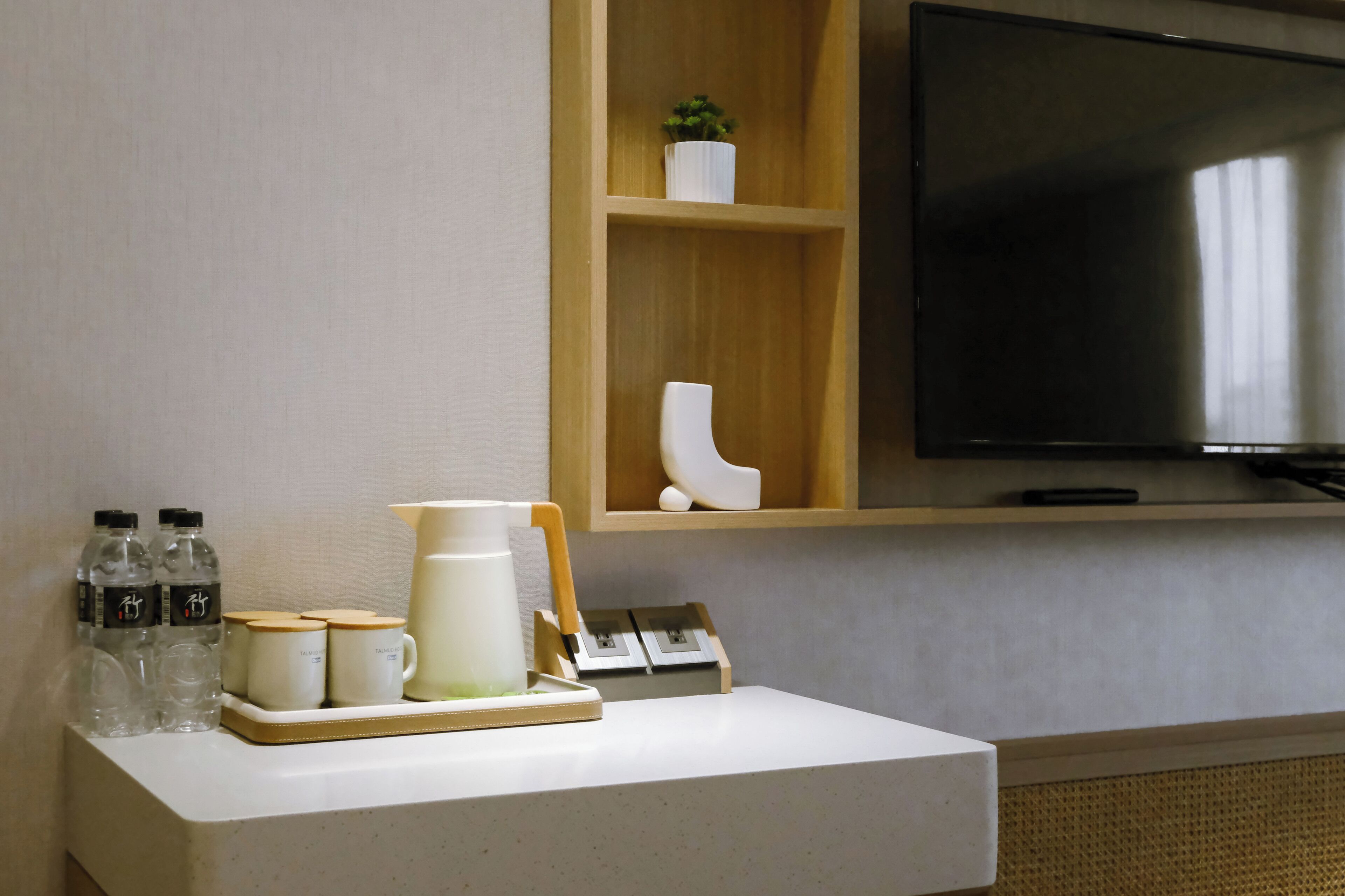 Une chambre d’hôtel équipée d’un téléviseur à écran plat fixé au mur, d’une petite étagère ornée d’objets décoratifs et d’une table sur laquelle se trouvent une carafe, des tasses et des bouteilles d’eau.