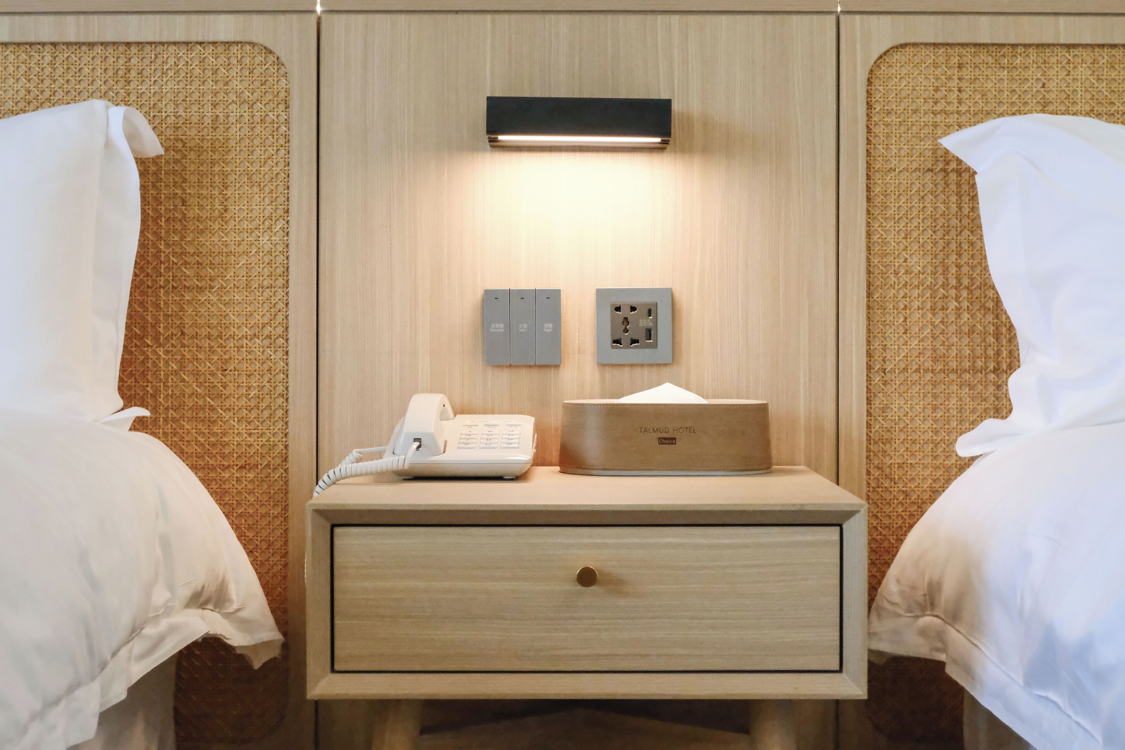 Een hotelkamer met twee bedden, een nachtkastje met een telefoon, een lamp en een doosje met het merk Talmud Hotel.