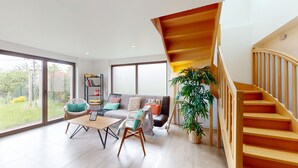A spacious living room with a wooden staircase, large windows, and a bookshelf.