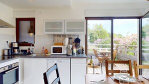 A modern kitchen with a dishwasher, oven, and coffee maker. There is a dining area with a table and chairs, and a view of trees and flowers through large windows.