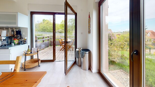 A modern kitchen with a dining area, large sliding glass doors to a deck, and a view of a garden and buildings.