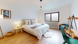 Un dormitorio con cama, mesitas de noche, lámpara colgante, ventana, radiador, cesta de mimbre y una silla.