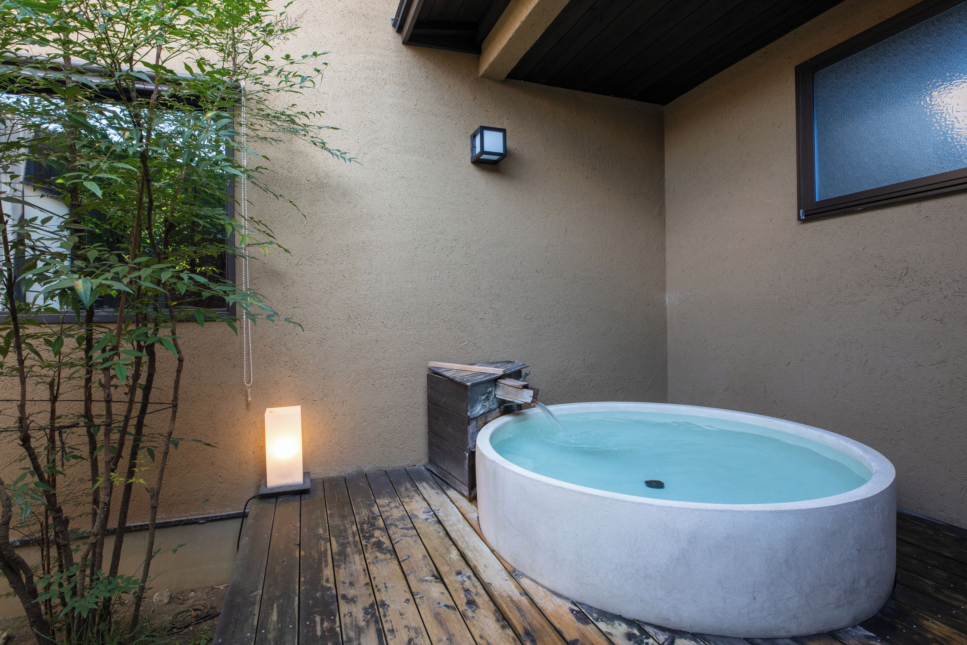 A hot tub in a covered outdoor area with a wooden deck, a wall-mounted light, and bamboo plants.