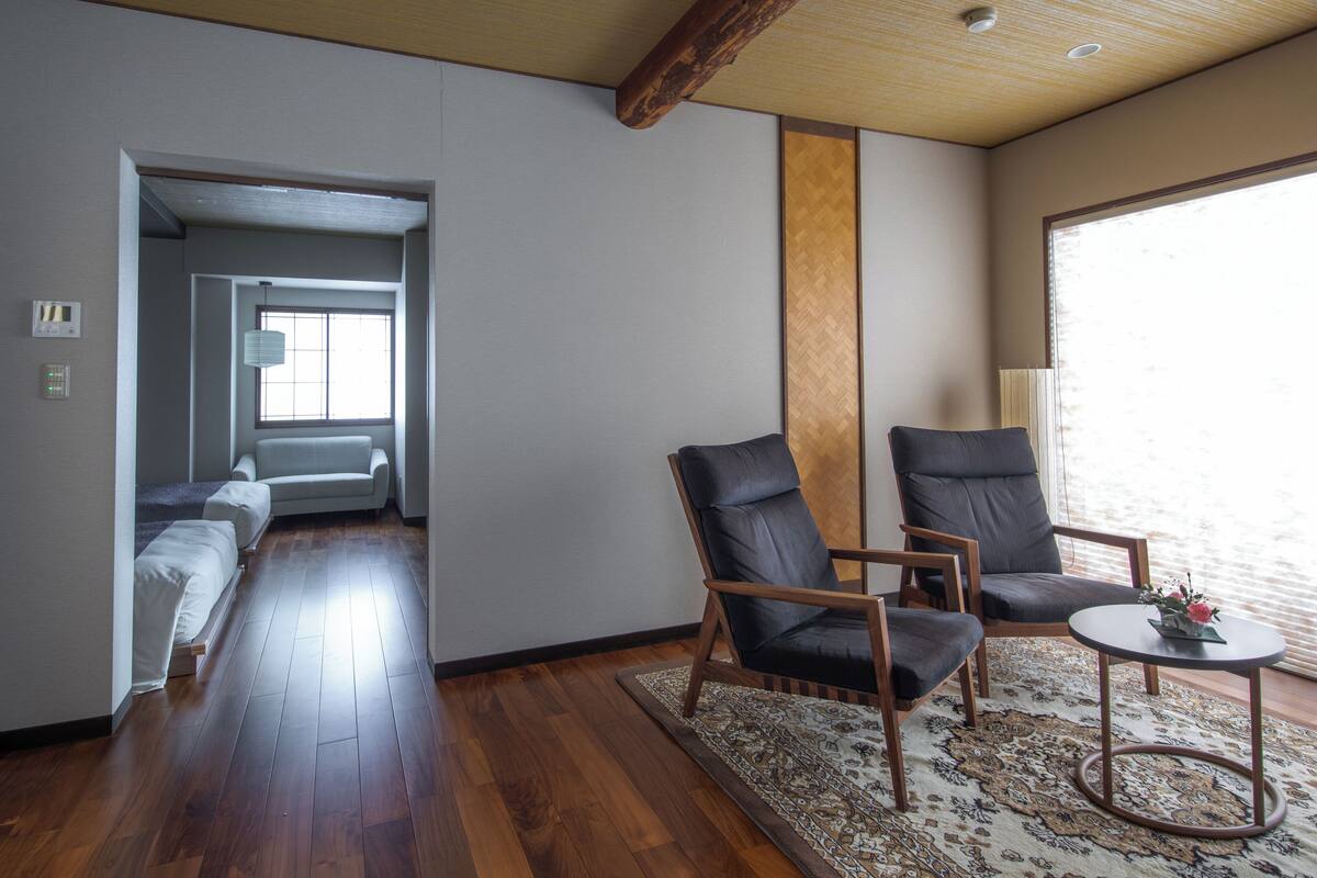 Ein modernes Wohnzimmer mit Holzboden, zwei Ledersesseln, einem kleinen runden Tisch und einem großen Fenster, das natürliches Licht hereinlässt.