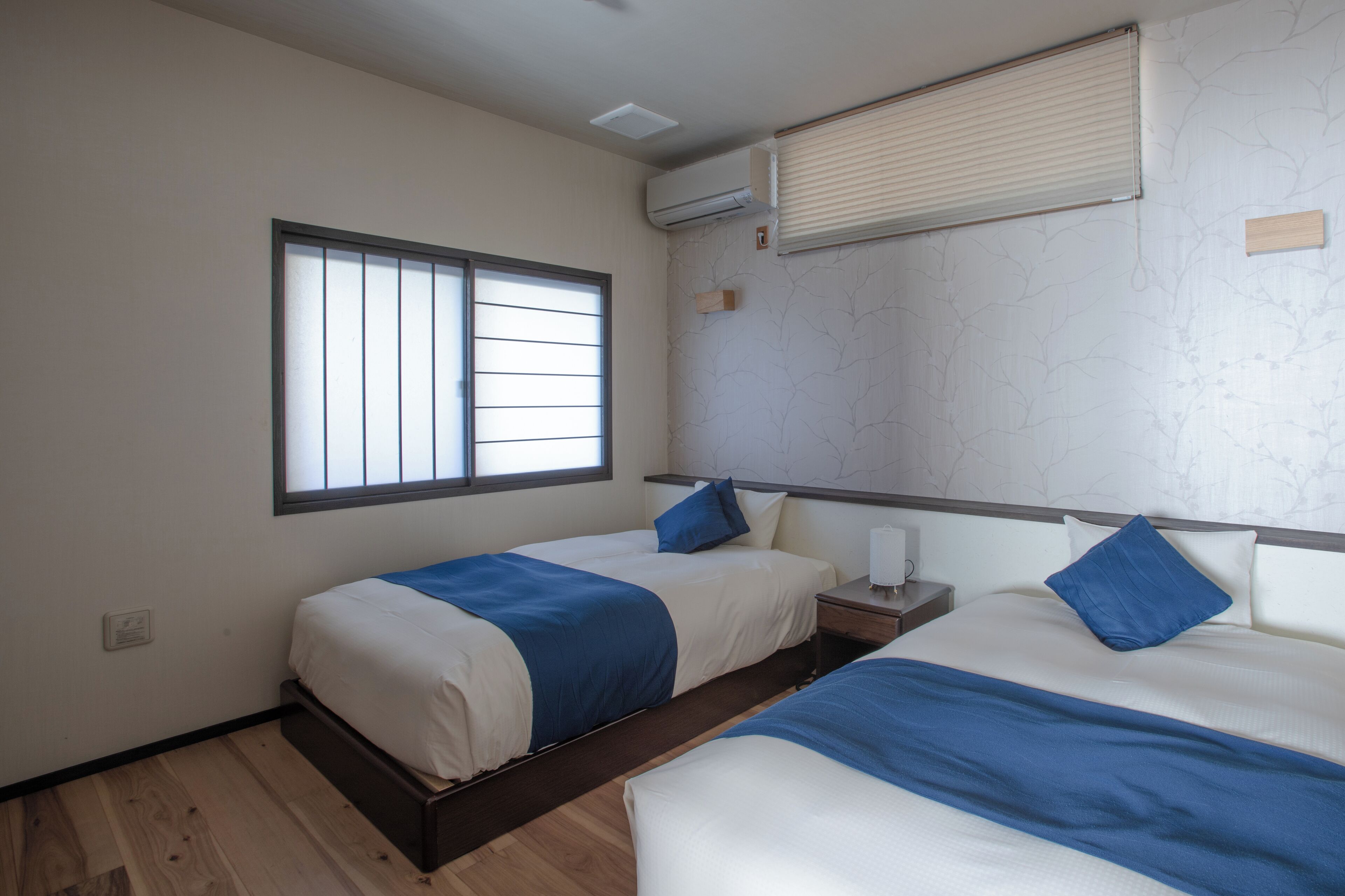 A hotel room with two beds, a window with blinds, an air conditioning unit, and a small bedside table with a lamp.