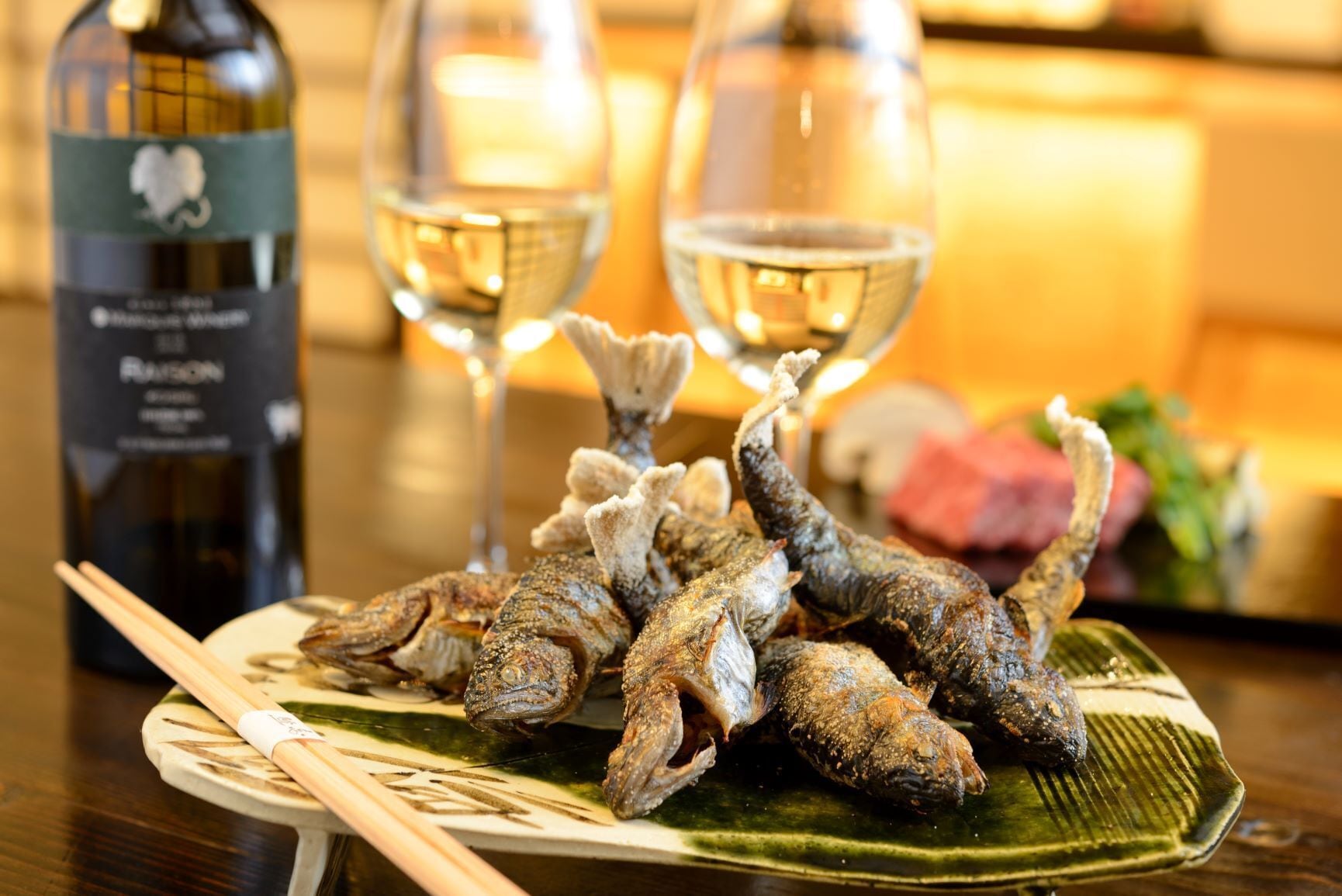 Une assiette de poisson frit, des baguettes et une bouteille de vin sur une table.