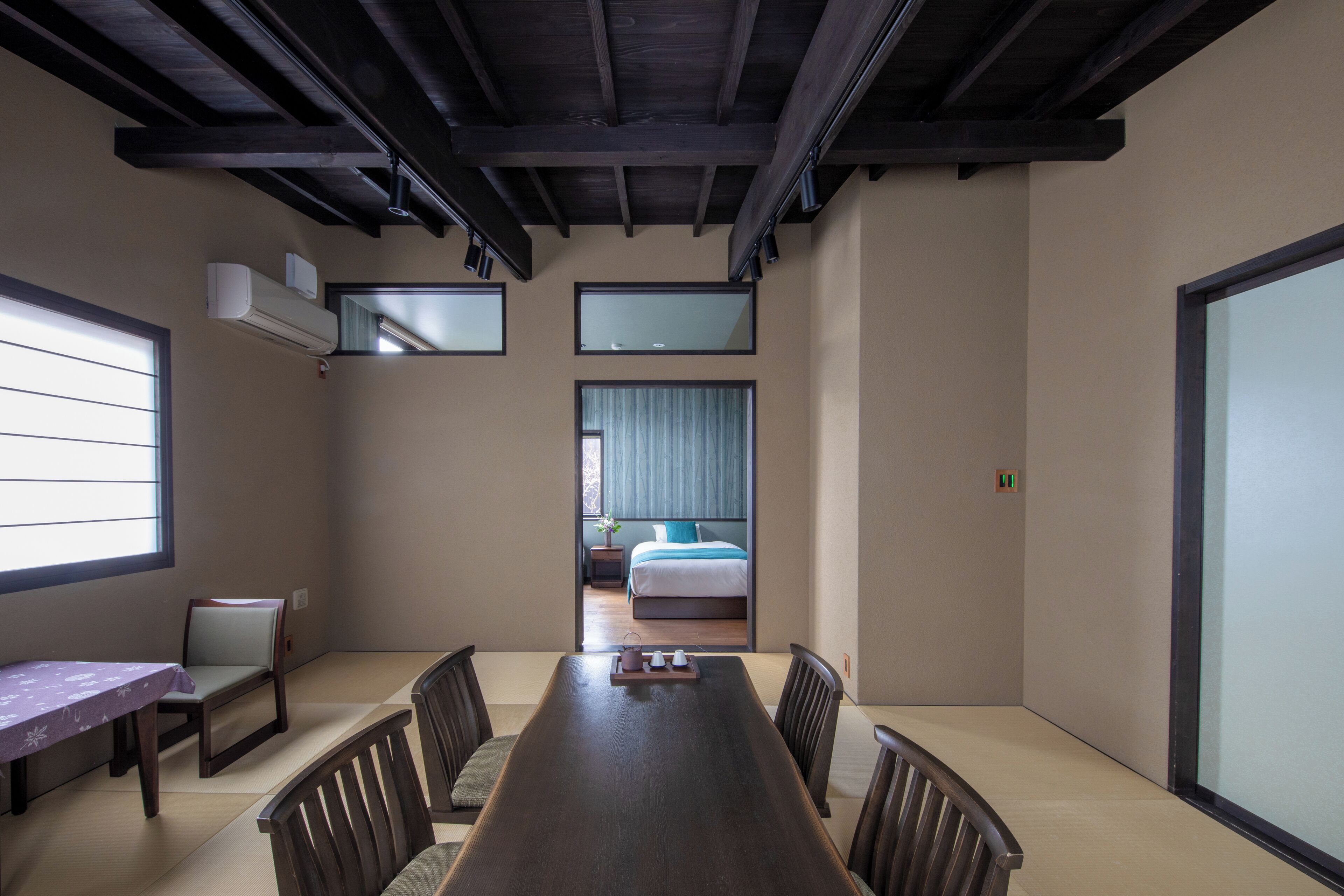 A hotel room with a large wooden dining table, a bed with blue curtains, and a view of a garden through a sliding door.