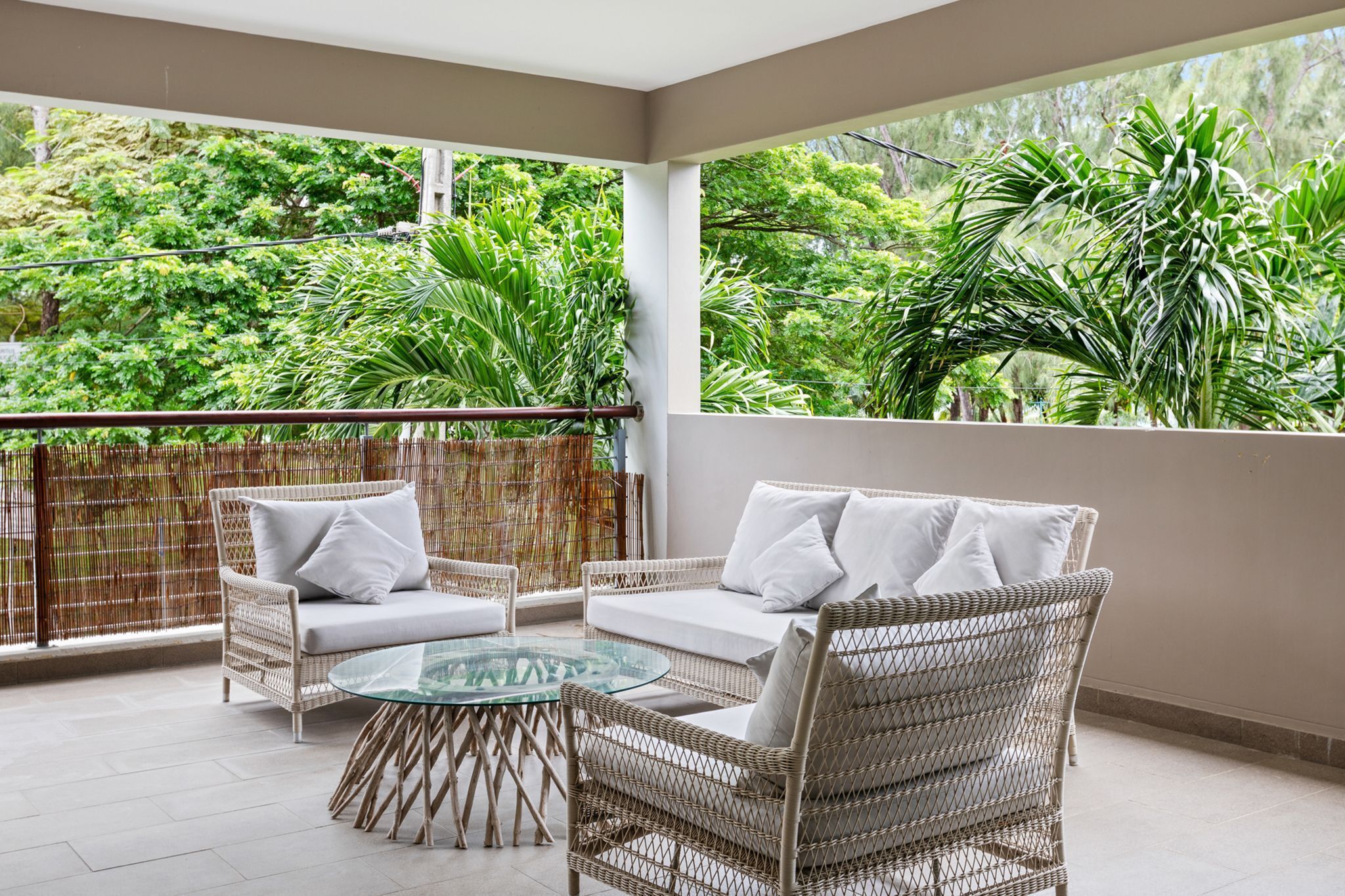 A balcony with wicker furniture, a glass table, and a view of lush greenery.