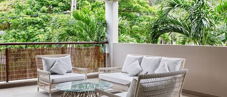 A balcony with wicker furniture, a glass table, and a view of lush greenery.