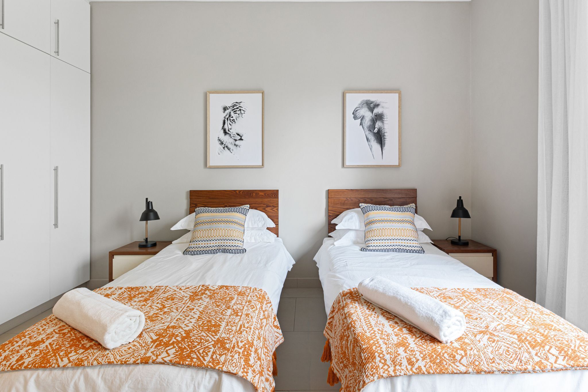 Une chambre d’hôtel avec deux lits, chacun recouvert d’une couvre-lit à motifs et agrémenté d’une serviette enroulée. Les murs sont ornés de tableaux encadrés, il y a une table de chevet avec une lampe et une armoire.