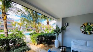 Uma sala de estar moderna com um sofá branco, uma mesa de centro e uma grande janela com vista para uma piscina e vegetação.